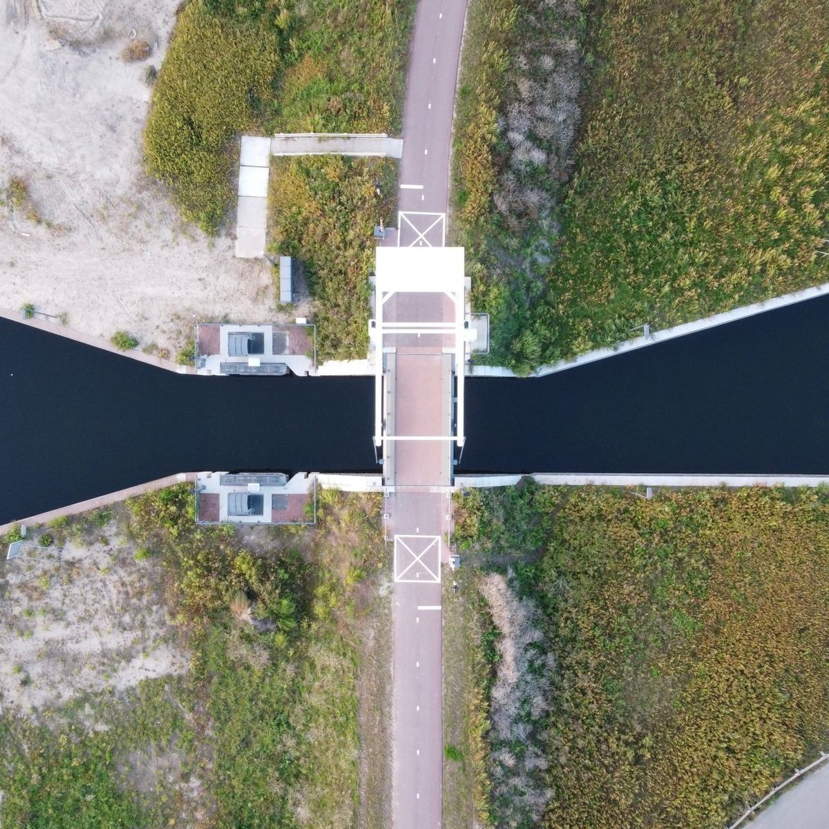 Luchtfoto recht van boven: een witte ophaalbrug over een donkere vaart, met fietspad en rietoevers